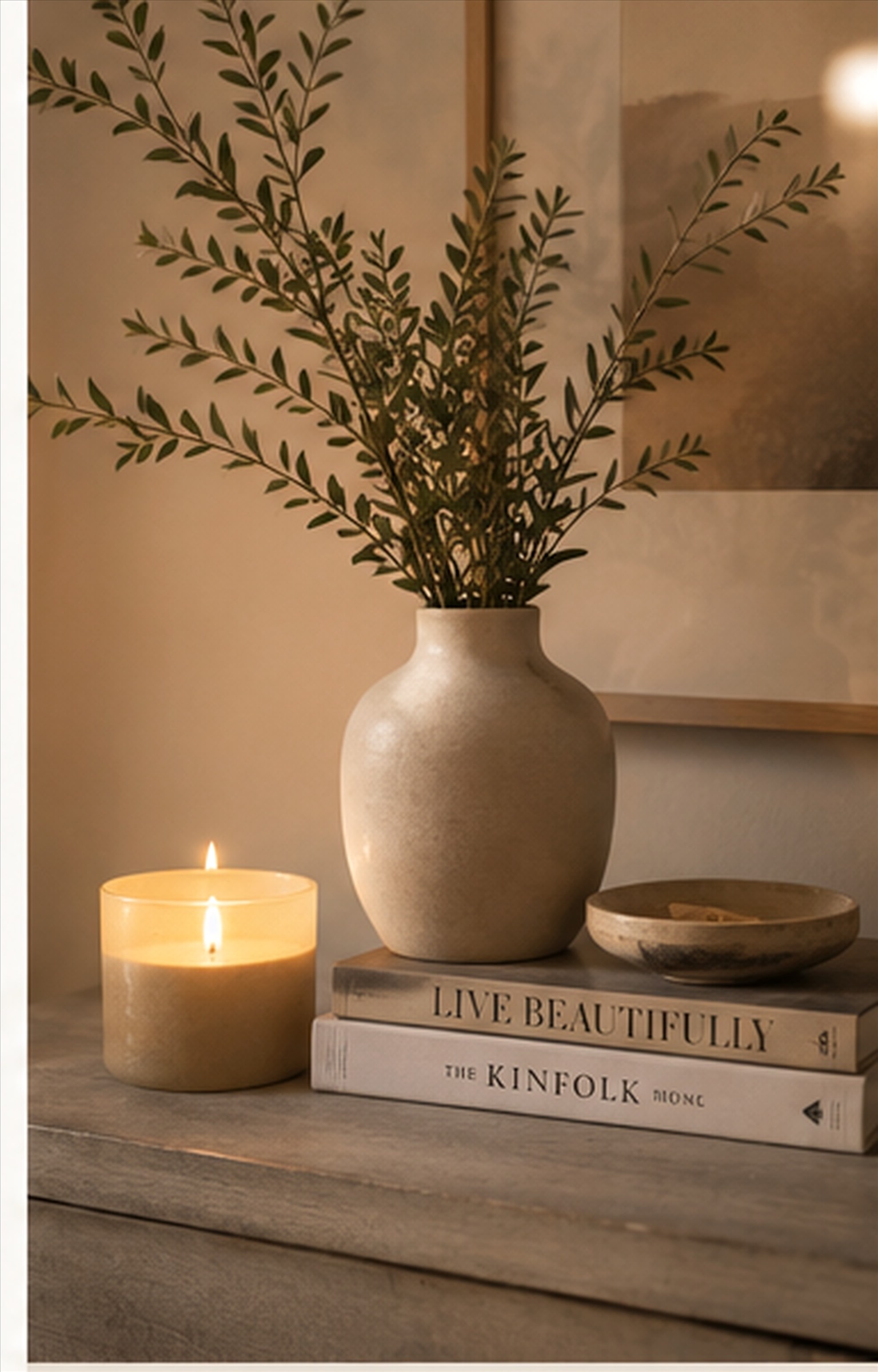 A styled bedside vignette with a lit candle, ceramic vase, and books stacked beneath fresh greenery.